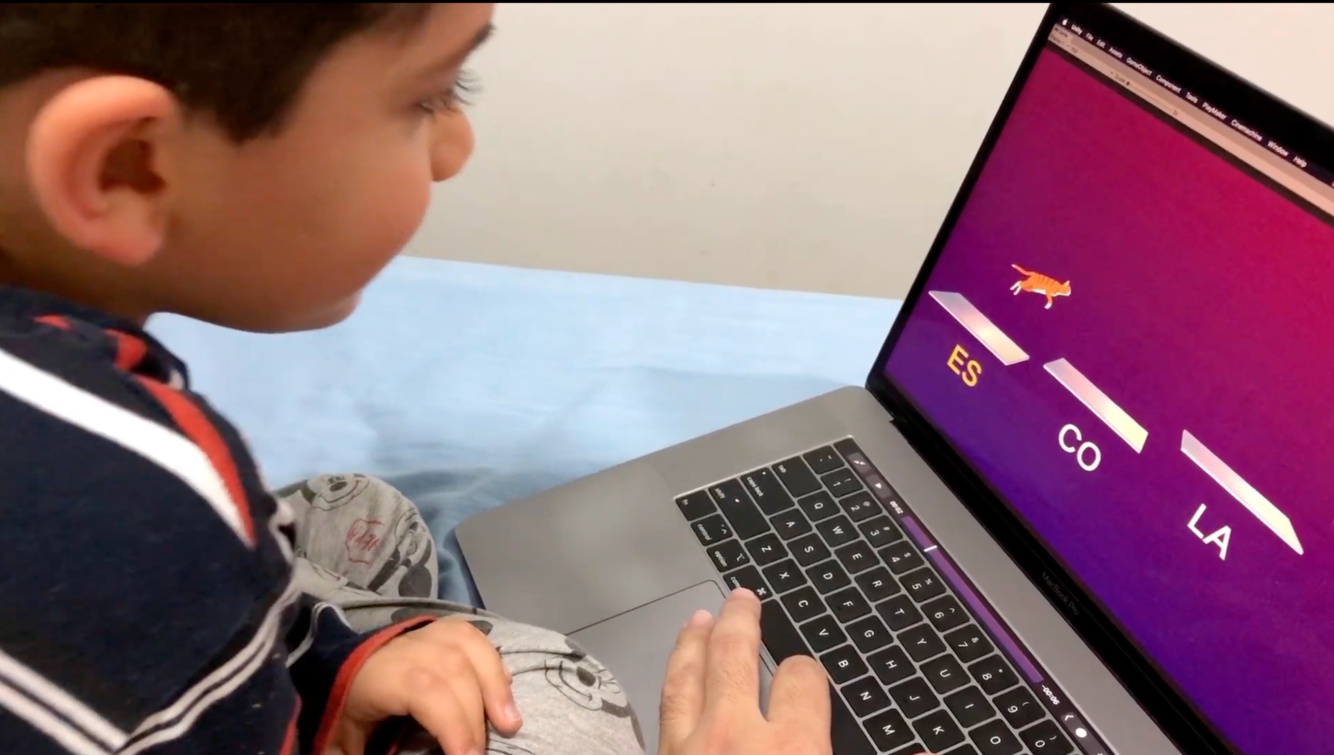 Miguel, de perfil, concentrado em frente ao notebook jogando uma versão inicial de 'Estoure o Balão' com a mão do pai apoiando no trackpad